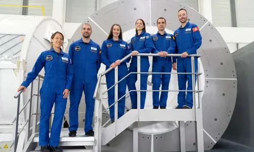 Katherine Bennell-Pegg standing on a staircase with her European Space Agency colleagues in blue jumpsuits and standing in front of space training equipment.