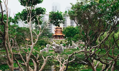 A Chinese-style pagoda sits in front of modern high rise buildings and is surrounded by trees in a garden